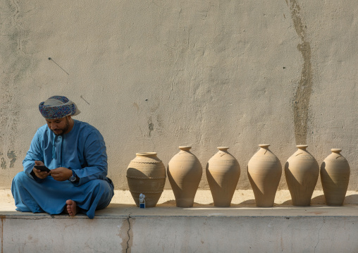 Omani man reading his mobile phone sit next to potteries, Ad Dakhiliyah Region, Nizwa, Oman