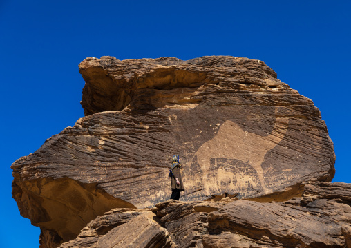Tourist standing in front of a life-sized camel petroglyph on a rock, Najran Province, Thar, Saudi Arabia