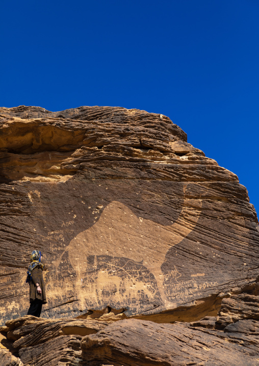 Tourist standing in front of a life-sized camel petroglyph on a rock, Najran Province, Thar, Saudi Arabia