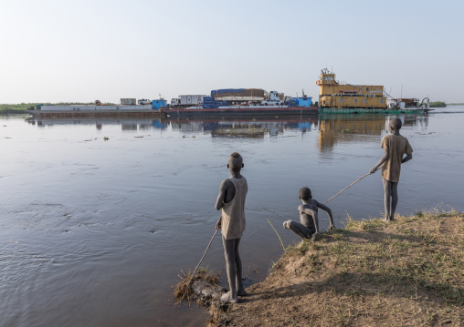 Barge passing in front of Mundari tribe boy fishing in river Nile, Central Equatoria, Terekeka, South Sudan