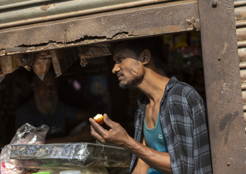 Bangladeshi man eating an apple below a shop iron curtain, Dhaka Division, Munshiganj, Bangladesh