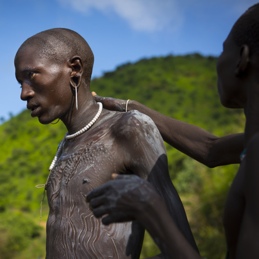 Shepherd from Suri tribe receiving help to decorate his body with camouflage paintings, Tulgit, Omo valley, Ethiopia